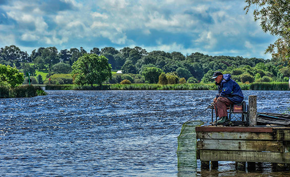 angler sitting on a wooden jetty fishing into a wide tree lined river on a sunny day/