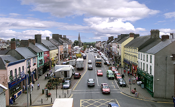 view down the main street of a busy town with cars parked either side of the road and brightly coloured buildings either side of the street/