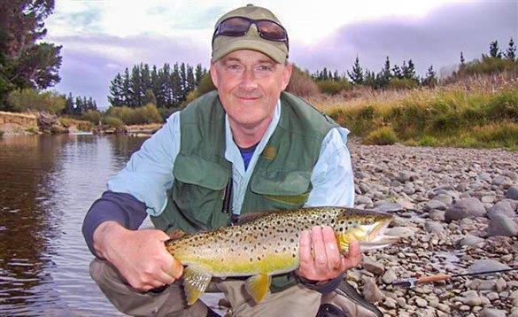 angler knelt beside a river holding a brown trout/