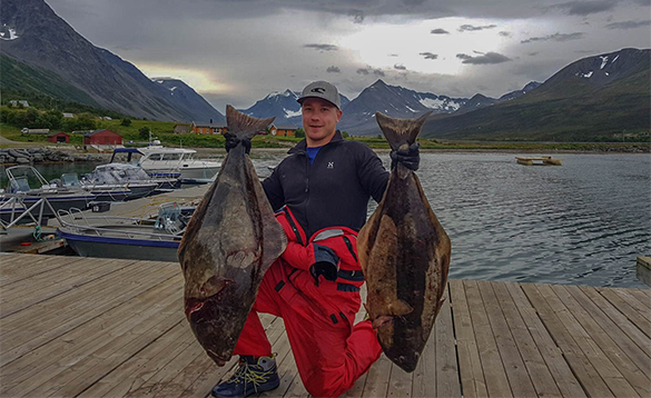 Angler on a pier holding two large halibut fish/