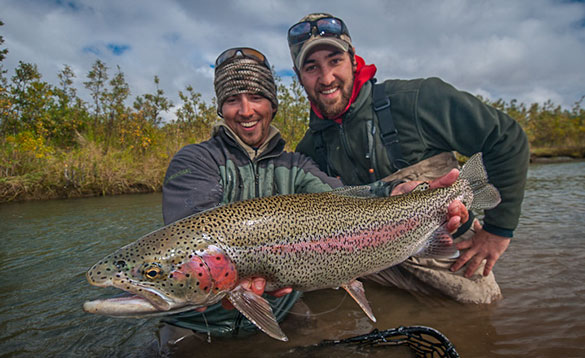 two anglers knelt in the river holding a large rainbow trout/
