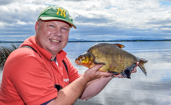 main in a red T shirt and green hat standing in front of a lake holding a bream/