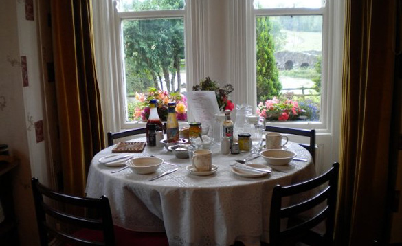 Round table in a bay window set for breakfast with views through the window to flower boxes and stone bridge in the bakcgorund