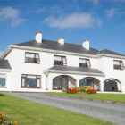 Gravel driveway leading to large white two storey house with lawns and colourful flower beds