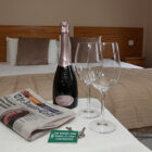 Table with champgane, two glasses and a newspaer on it next to a double bed