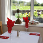 Table next to a window with white table cloth and red napkins and view of sheep in a field