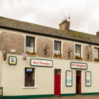 Two storey Irish pub with cream painted lower half with red door and four lights between the upper room windows