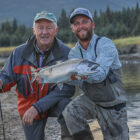 Two anglers beside a river - one holding a fishing rod and the other holding a coho salmon