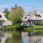 View across a lake to thatched dorma bungalows with lawn gardens and trees