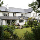 Large two-storey white guest house with a grey slate roof, featuring a manicured lawn, lush gardens, and a classic front porch.