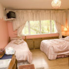 Cosy pink-themed bedroom with a double bed and a single bed featuring matching pink striped bedding, large windows with cream ruffled curtains, and a sparkling pink chandelier.