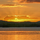 Vibrant orange and gold sunset over a calm lake, with the sun's rays breaking through a cloud and illuminating the silhouette of distant hills.