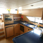 Modern galley kitchen inside a cruiser boat, featuring light wood cabinets, blue countertops, a small stovetop, sink, and a fire extinguisher mounted on the wall.