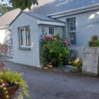 Light grey cottage exterior with a small porch, surrounded by lush pink hydrangeas and green bushes, with a hanging flower basket in the foreground.