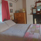 Traditional cottage bedroom featuring a double bed with a patchwork quilt, a vintage fireplace with rose-patterned tiles, and a large wooden dresser.