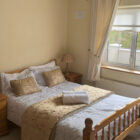 Bedroom with pine double bed infront of a window with white linen and gold cushions and throw