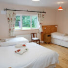 Spacious triple bedroom with light wood flooring, featuring one double bed and two single beds with white linens and a wide window overlooking a garden.