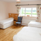 Spacious triple bedroom with light wood flooring, featuring one double bed and two single beds with white linens, a blue floral armchair, and a wide window overlooking a green field.
