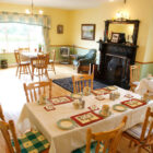 Spacious traditional dining and sitting room featuring light yellow walls, wooden flooring, several dining tables with white linens, a dark ornate fireplace, and green leather sofas.