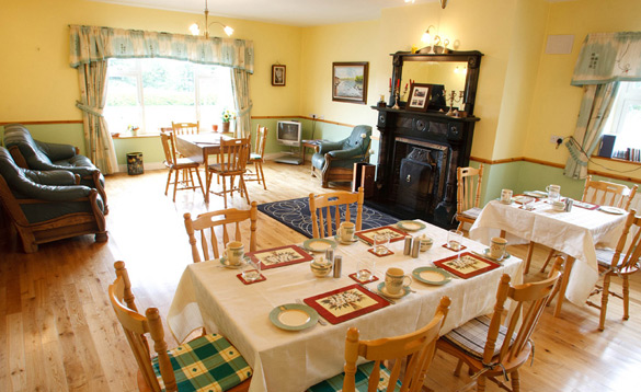 Spacious traditional dining and sitting room featuring light yellow walls, wooden flooring, several dining tables with white linens, a dark ornate fireplace, and green leather sofas.