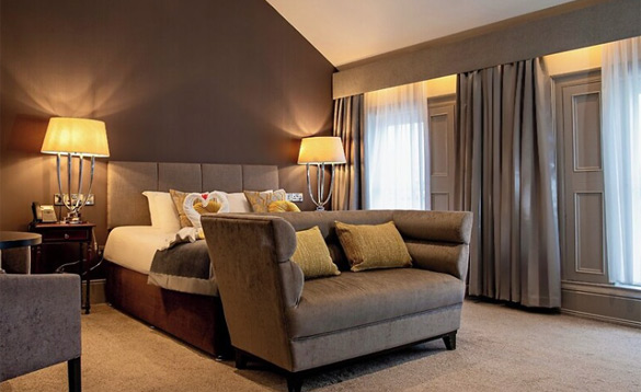 A luxury hotel room with a plush grey sofa, a king-size bed featuring swan-folded towels, and elegant warm lighting.
