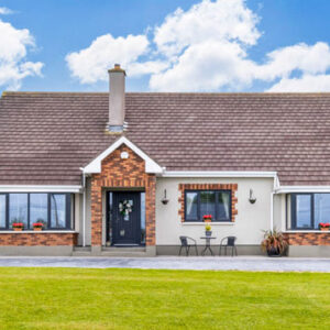 Front view of a modern bungalow with a light grey facade, red brick accents around the windows and porch, a dark tiled roof, and a large green lawn under a blue sky.