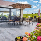 Spacious modern grey stone patio with a round outdoor dining table, black ornate chairs, and a large tan umbrella, overlooking a green field under a blue sky.