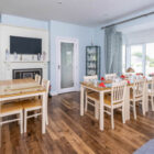 Bright, modern dining room with light blue walls, featuring two light wood dining tables, a fireplace with a mounted TV, and a large bay window with grey patterned curtains.