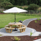 Landscaped garden with circular stone patio, wooden picnic table, white parasol, and manicured lawn.