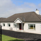 Modern white bungalow with red accents, large windows, and a black tarmac driveway in a rural setting under a blue sky