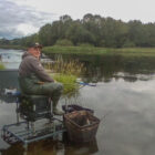 An angler in green waders sitting on a specialized fishing platform in a calm lake, surrounded by reeds and a dense forest background.