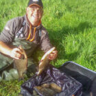 A smiling fisherman in green waders sitting in a grassy field, holding two large fish over a full fishing net.