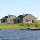 Two identical grey pebble-dash bungalows with slate roofs on a grassy hill overlooking a lake with a small wooden jetty and boat.