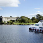 A white power cruiser with a blue canopy navigating a wide river toward a large, historic stone warehouse building on the grassy bank under a clear sky.