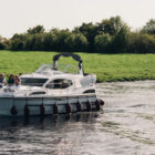 A group of people relaxing on the bow of a white cruiser boat as it travels down a calm river past green grassy banks.