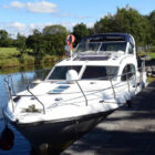 A sleek white power cruiser boat moored at a stone dock on a calm waterway, with lush green trees and a clear blue sky in the background.