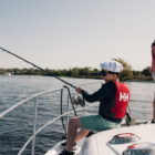 A man and a young boy wearing a sailor's hat and red life jacket fishing from the deck of a white boat on a wide, calm river under a clear sky.