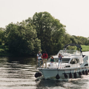 A group of four people wearing life jackets standing on the bow of a white cruiser boat as it travels down a calm river lined with lush green trees.