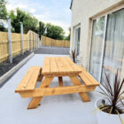 A wooden picnic table on a paved patio area next to a cream-colored cottage, featuring a modern wooden fence and a potted plant.