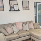 A modern grey corner sofa with decorative striped and pink cushions, situated under three abstract framed art pieces on a white wall.