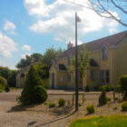 A row of two-storey yellow townhouses with grey roofs and blue window trim, situated behind a gravel driveway with small shrubs and a green lawn under a sunny sky.