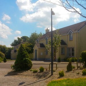 A row of two-storey yellow townhouses with grey roofs and blue window trim, situated behind a gravel driveway with small shrubs and a green lawn under a sunny sky.