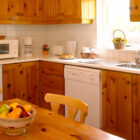 A rustic country kitchen with pine cabinetry, white tiled walls, a fruit bowl on a wooden table, and various white appliances including a microwave and dishwasher.