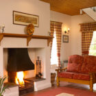 A warm, traditional living room featuring a large white fireplace with a roaring fire, a wooden mantel, and a red patterned sofa set against white walls and a wooden ceiling.