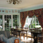 Dining room with two dark wood tables and eight chairs, large windows with red multi patterned swag curtains