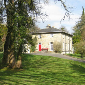 Drive leading up a grassy hill towards a large two storey stone house with red door and grey tiled roof