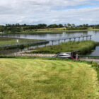 A metal fishing pier extending into a calm lake, with a small utility building and a parked car on the grassy shore under a cloudy sky.