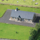 An aerial view of a stone-fronted cottage with a grey slate roof, surrounded by a paved driveway and large green lawns, with sheep grazing in a nearby field.