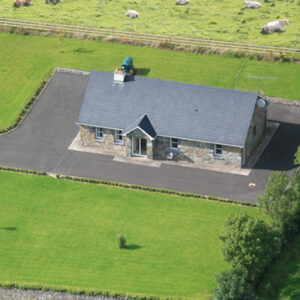 An aerial view of a stone-fronted cottage with a grey slate roof, surrounded by a paved driveway and large green lawns, with sheep grazing in a nearby field.