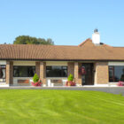 Stone bungalow with wooden benches outside and lawn and gardens to the front